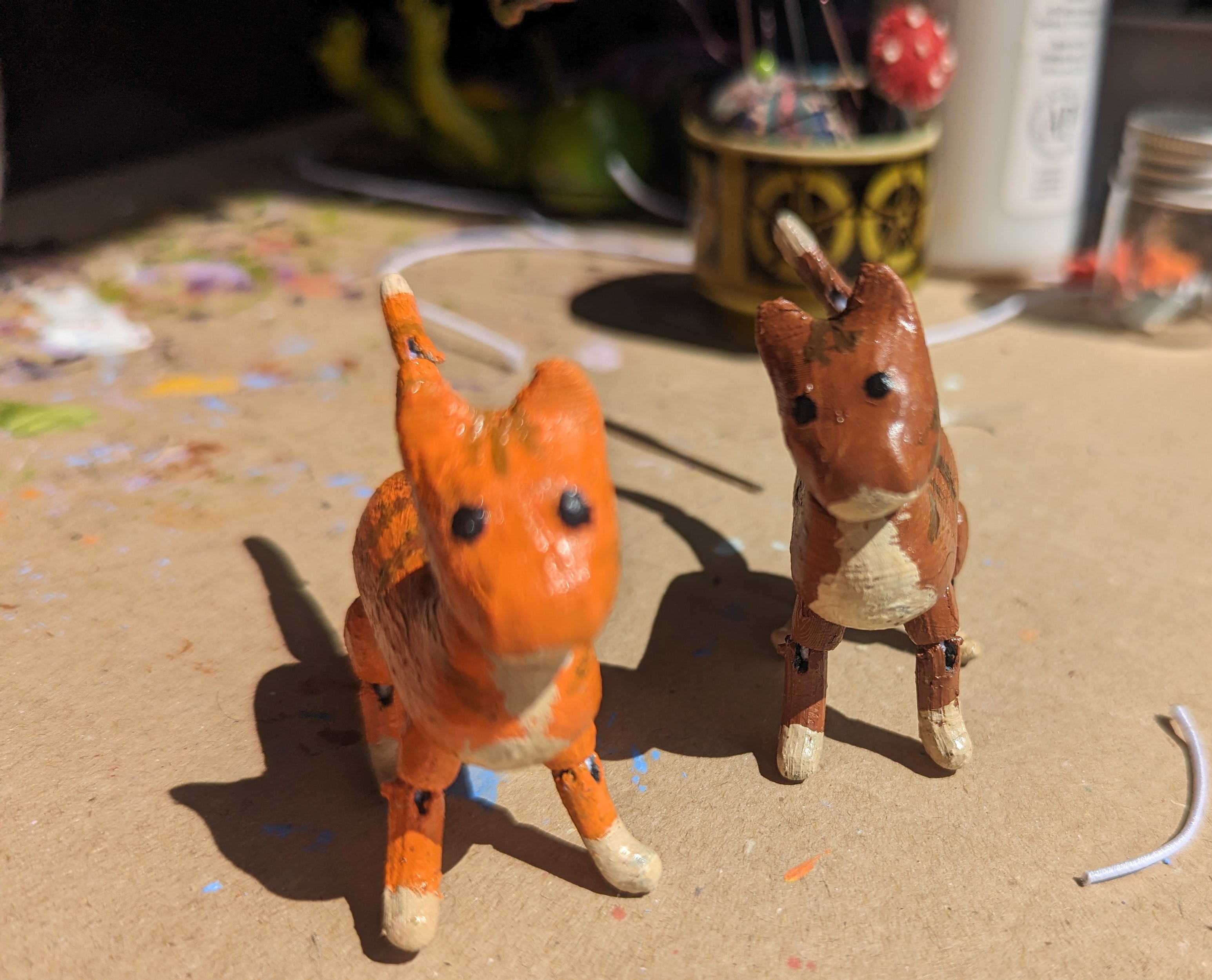 A pair of articulated cat dolls on a cluttered worktable, one painted tabby and one ginger. They're looking toward the camera.