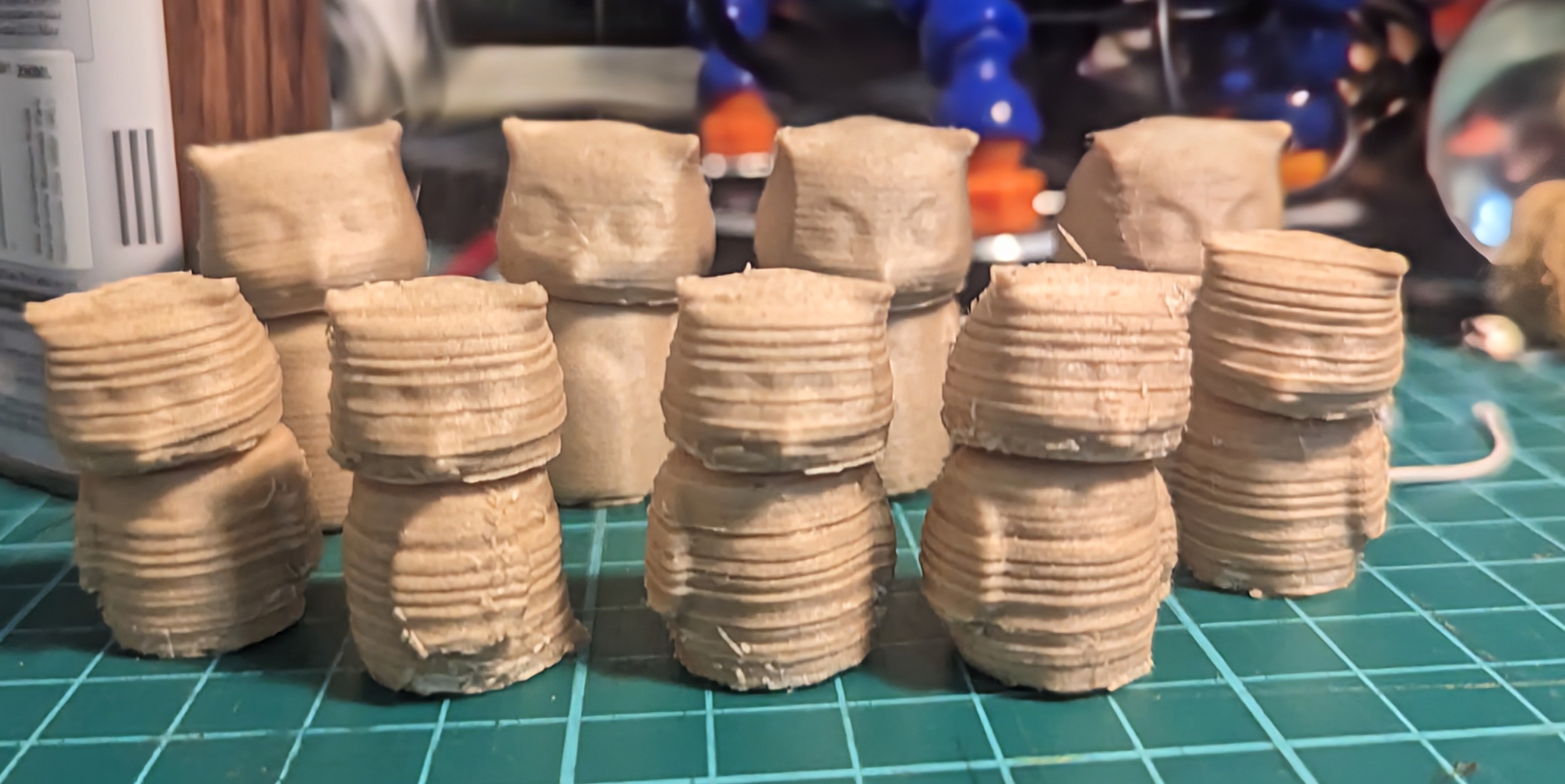 Two rows of 3d printed owls on a cluttered, green work mat. There are five in the front row that are noticably rougher, with pronounced horizontal lines and splinter-like imperfections. The four in the back are larger and almost completely smooth.