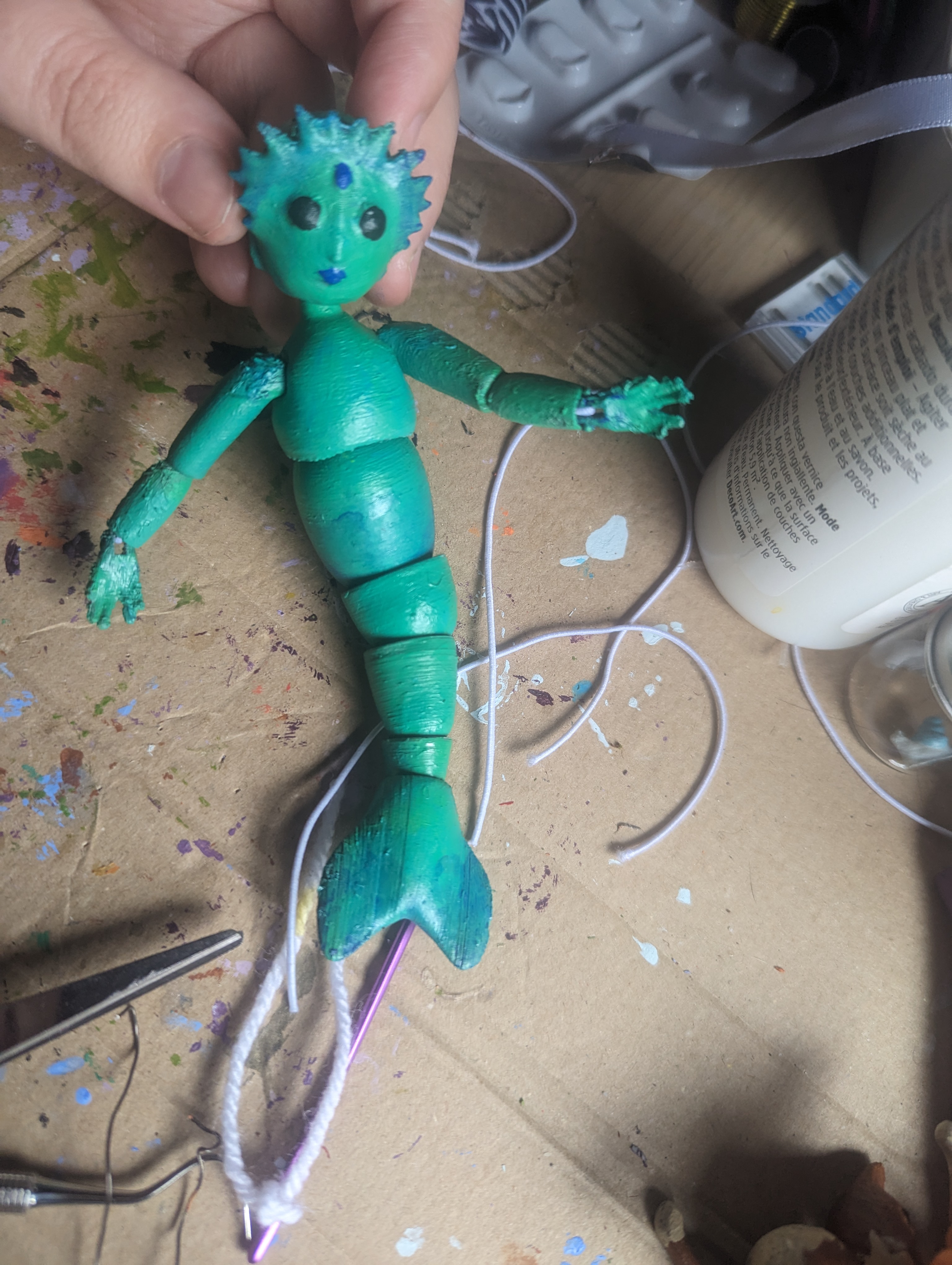 A seafoam green mermaid doll with a crown of fins looking toward the camera. It has four joints in its tail that let it twist smoothly.