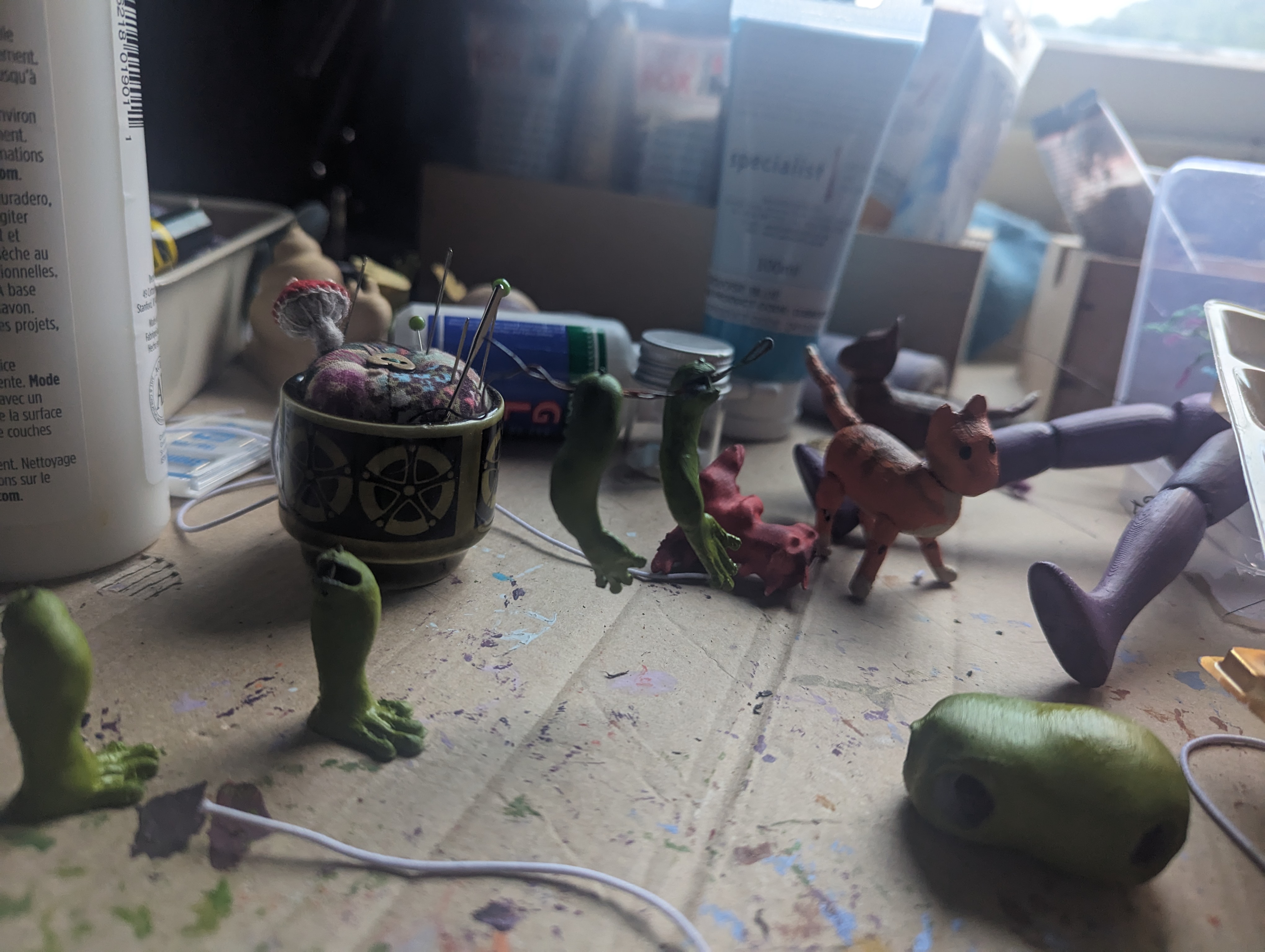 A slightly fuzzy picture of a cluttered worktable covered in various paint stains and painted figures. Two frog-like doll arms are hanging from a wire in the foreground.