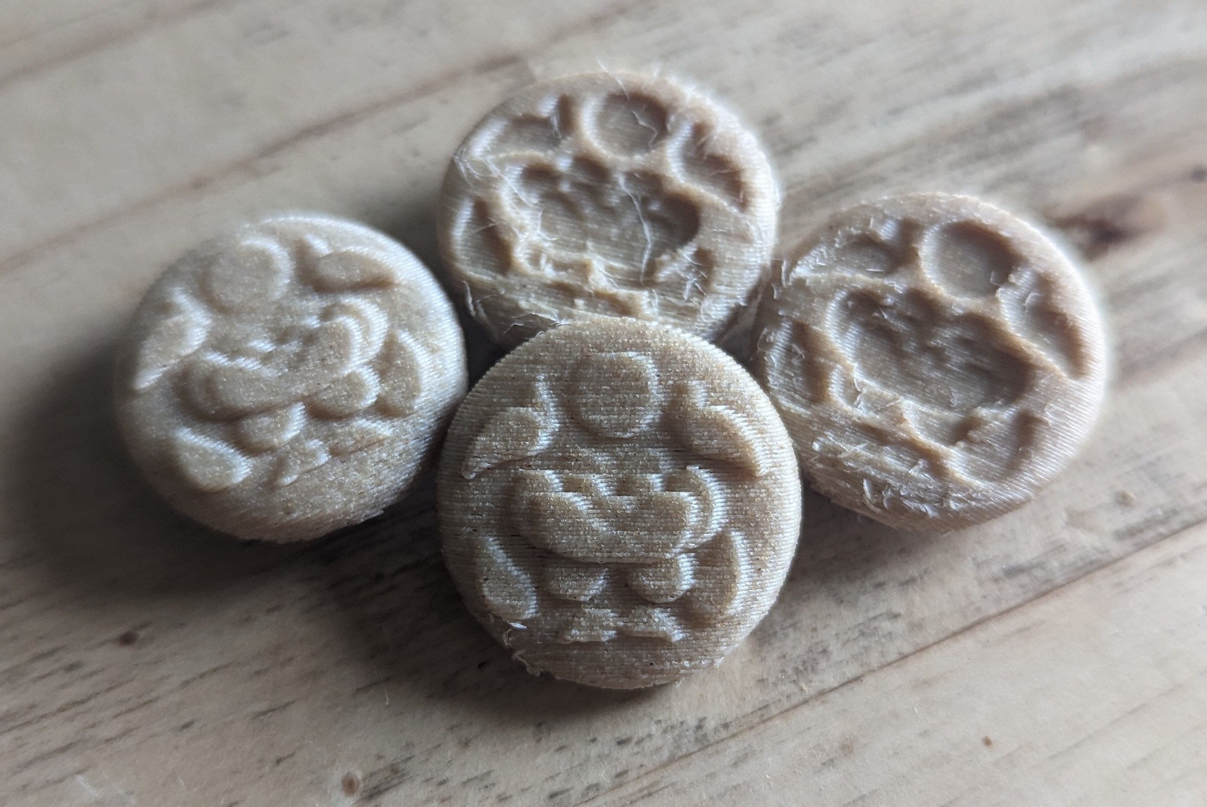 A photograph of four buttons 3d printed with wood filament on a wooden table. They all have a design of a lotus surrounded by leaves under a disk representing the sun or moon. Two of them have it potruding out in layers and two have it inverted inwards. The inverted buttons have more stringing and imperfection that look like splinters.