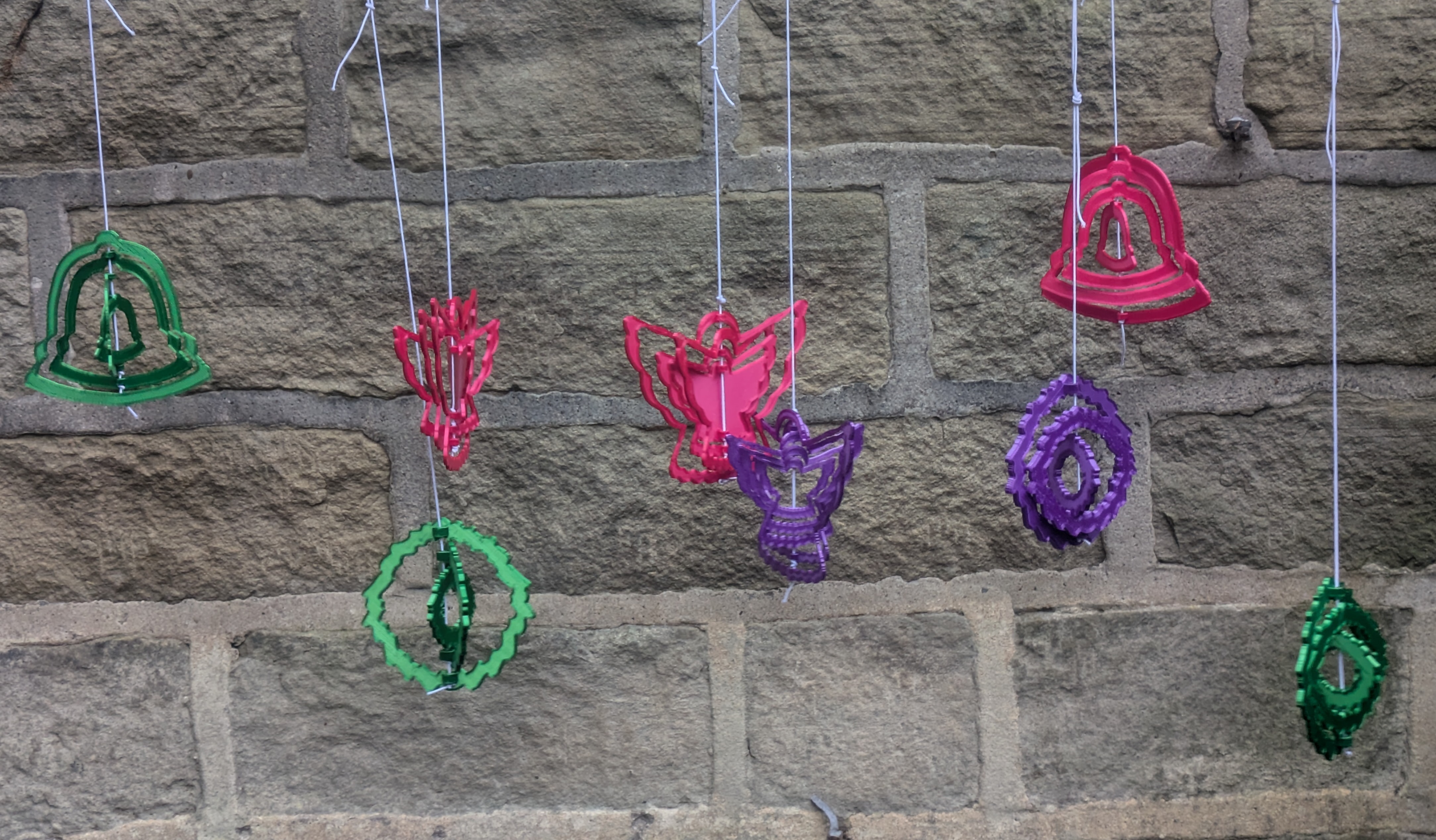 A set of multicoloured christmas ornaments hanging in front of a brick wall, consisting of concentric rings in various shapes.