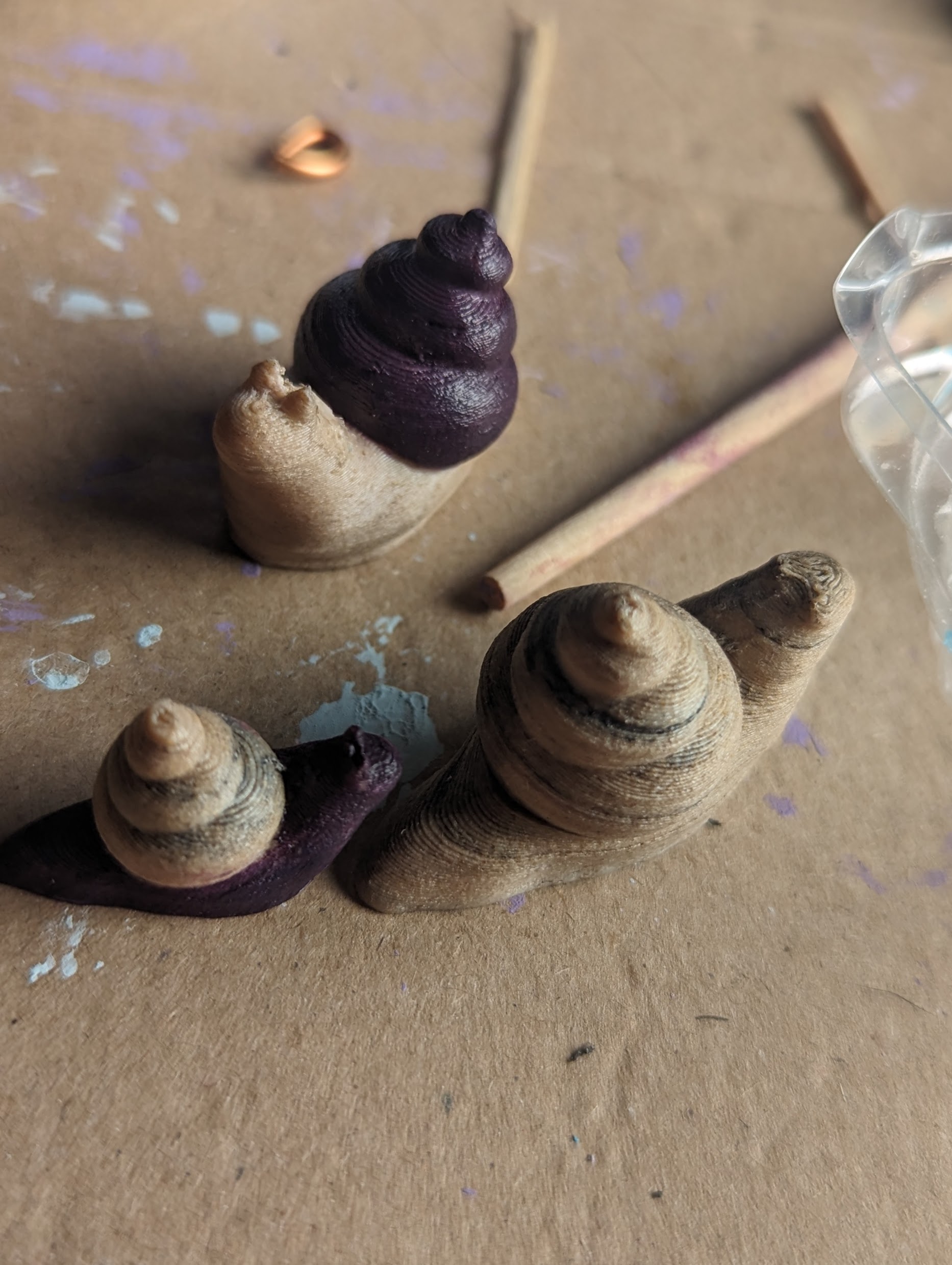 Three 3d-printed snails on a paint-spattered surface. They have been printed in wood-mix filament and have a wood-like appearance One has its body painted purple and is significantly smaller and has a purple shell.