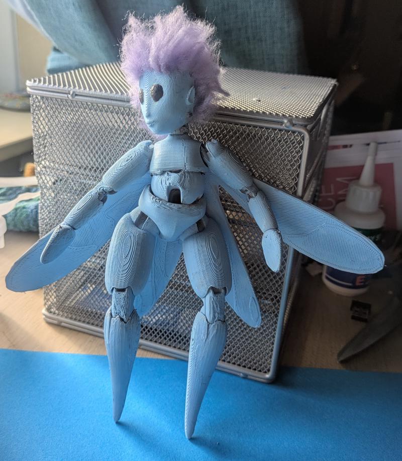 A fairy doll standing on a blue table. It is about 30 cm tall, similar to a Barbie and blue with purple hair.