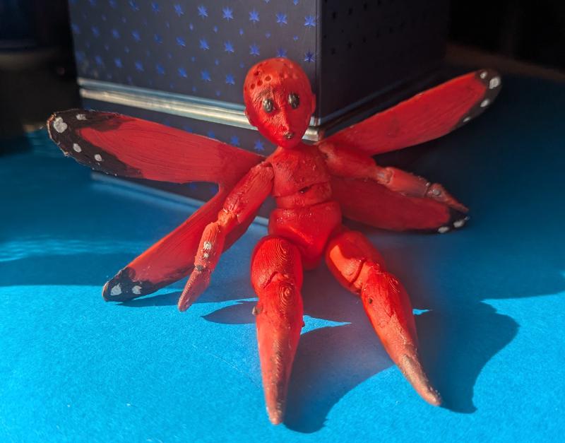 A fairy doll sitting on a blue table. It is about 15cm tall, about half the size of a Barbie, and orange, without any hair, but painted like a monarch butterfly.