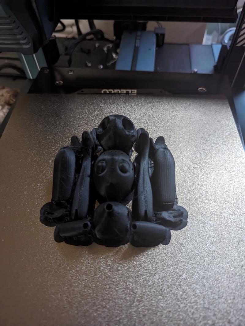 The bed of a 3D printer with the parts of the above frog doll laid out in black plastic.