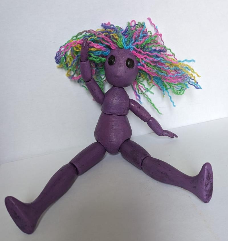 A humanoid doll painted purple with wild rainbow hair sitting down on a white background.
