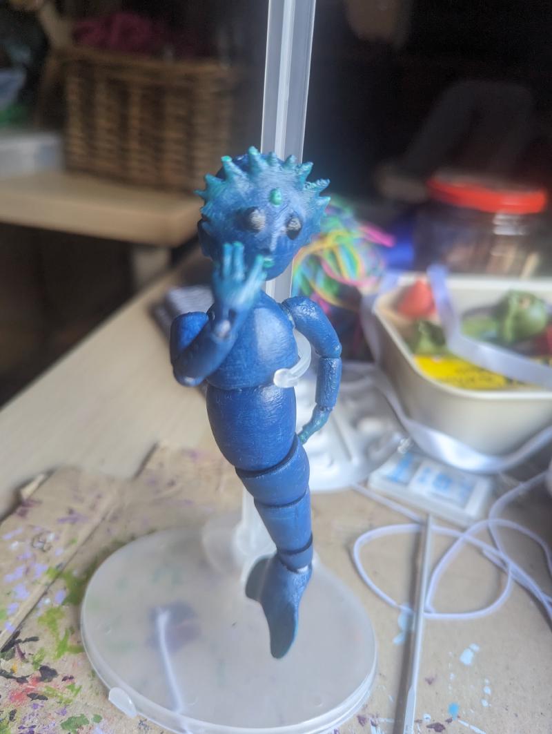 A blue mermaid doll on a doll stand in a cluttered workshop. It has its right hand dramatically coving its face and its left hand on its hip.
