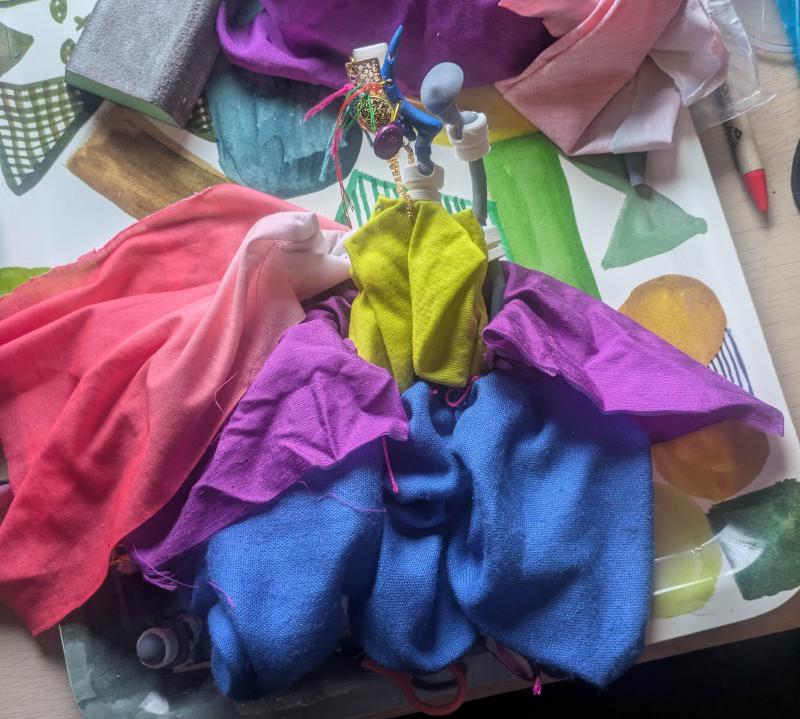 A photograph of the grey and white jointed skeleton, in a roughly humanoid shape, lying on a table with a costume loosely draped over it. it has a pale yellow shirt, a purple overdress, voluminous blue trousers and a cape with a peach gradient.